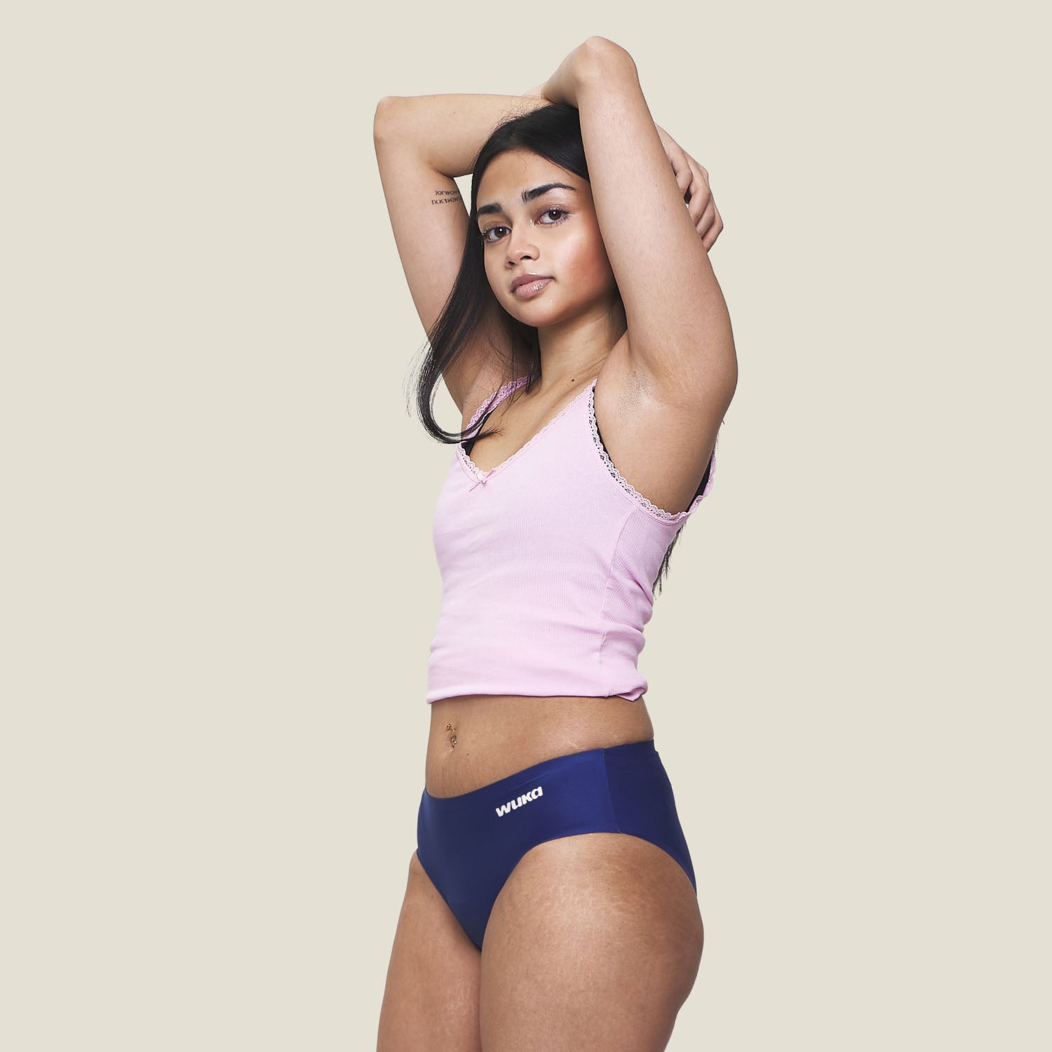 Teen girl wearing WUKA Teen Stretch Seamless Period Pants in blue for heavy flow, hands raised above head, showcasing full front view against a beige background.