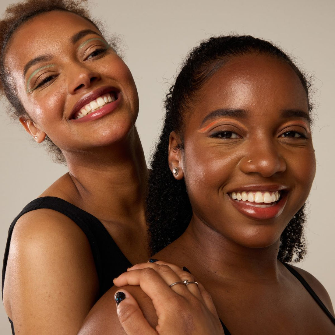 Two smiling women with bold eye makeup and glowing skin, standing close in black tops—celebrating sisterhood, confidence, and period-positive energy.