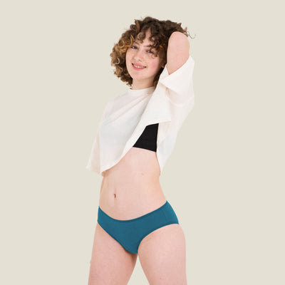 Teen girl wearing WUKA Teen Swim Bikini Brief in blue for light period flow, styled with a loose white top, posing confidently with hand in hair on a neutral background.