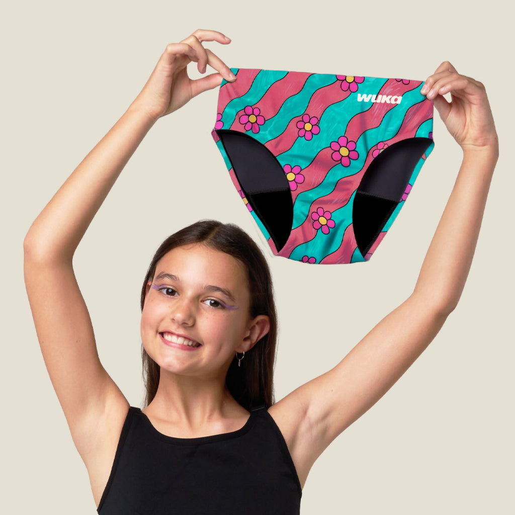 A young girl with long brown hair holds up a pair of colourful midi brief period pant bottoms above her head against a solid light beige background. The midi brief feature a vibrant pattern of pink and teal wavy stripes with purple flowers, black side panels, and a white 'WUKA' brand logo on the front right hip.