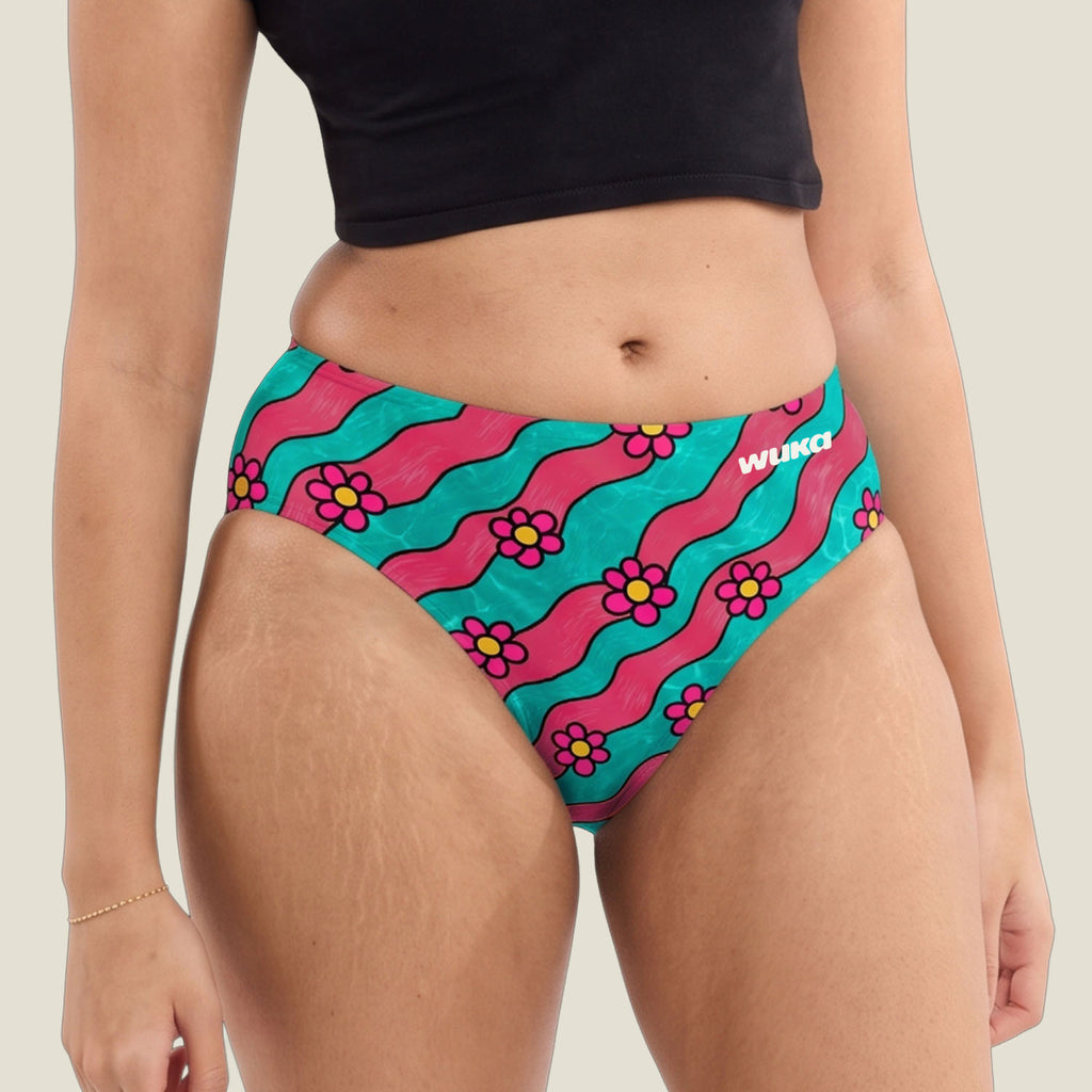 A close-up of a model wearing midi brief period pant bottoms with a retro pattern of wavy coral pink and teal stripes, accented with pink and yellow flowers. The brand name 'WUKA' is printed in white on the right hip.